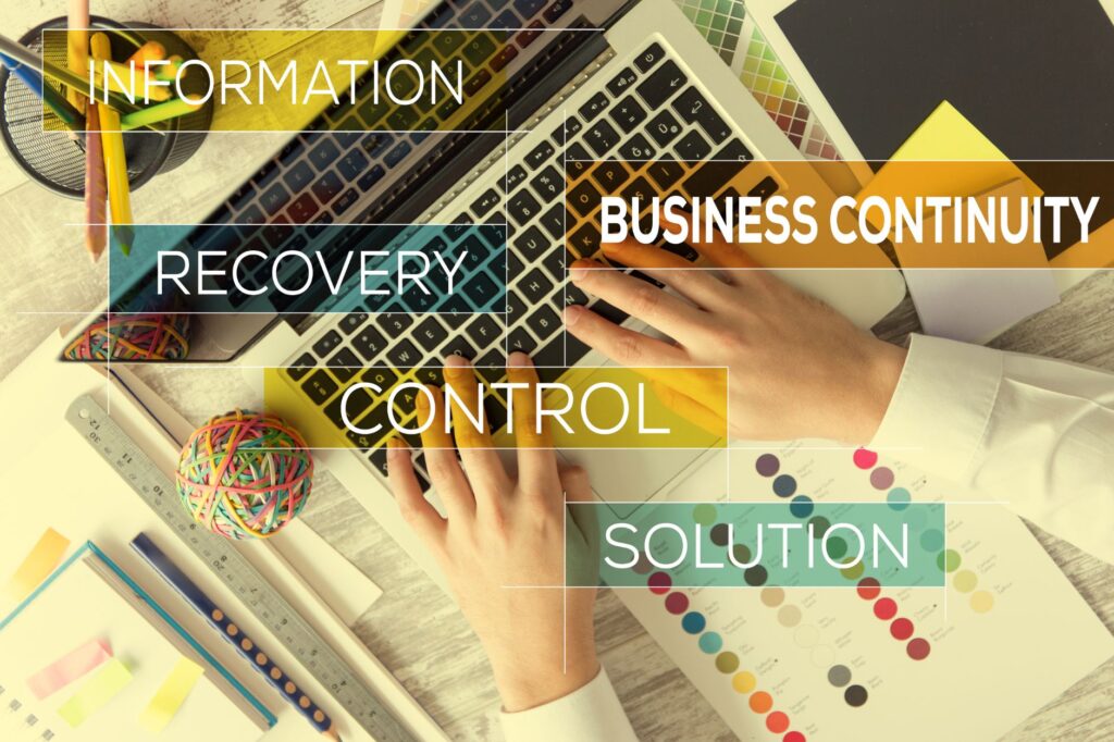 Hands typing on a laptop keyboard with text overlays: "Information," "Recovery," "Business Continuity," and "Control." Vibrant office supplies add a splash of color, emphasizing the seamless integration of technology and creativity in ensuring uninterrupted business operations.